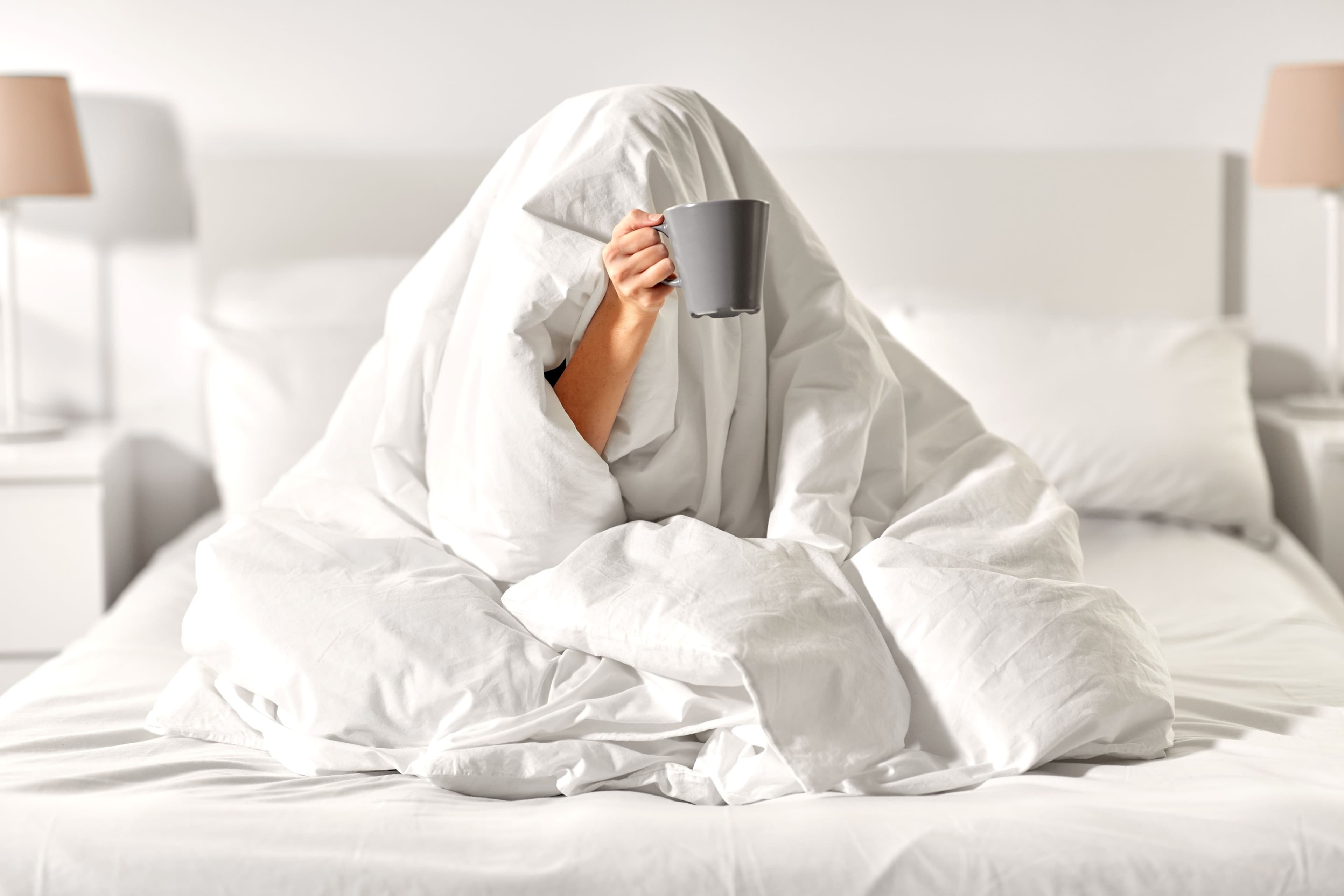 Person wrapped head to toe in a white doona holding a grey mug