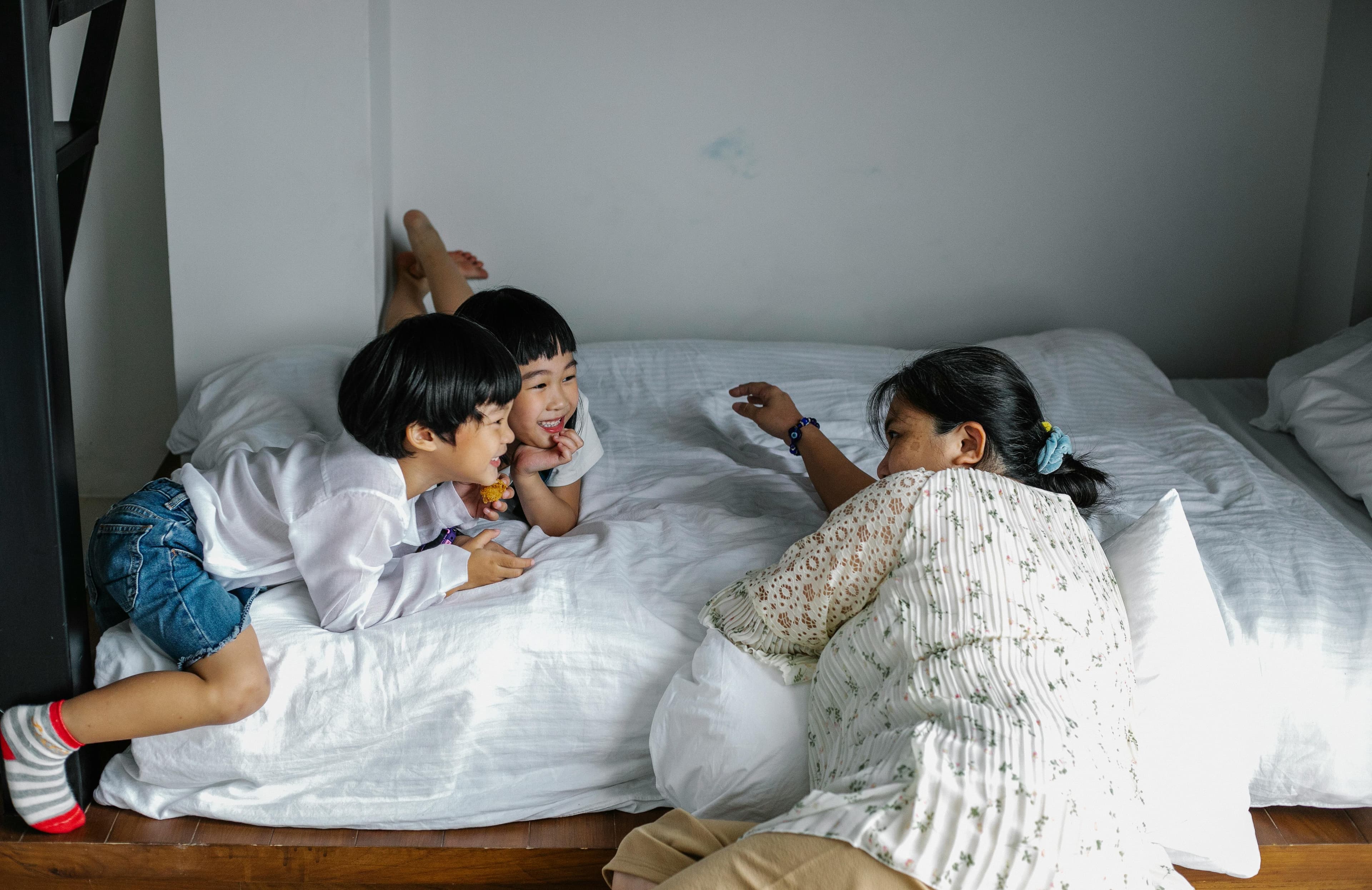 Mother chatting with two laughing young children on a white-sheeted bed