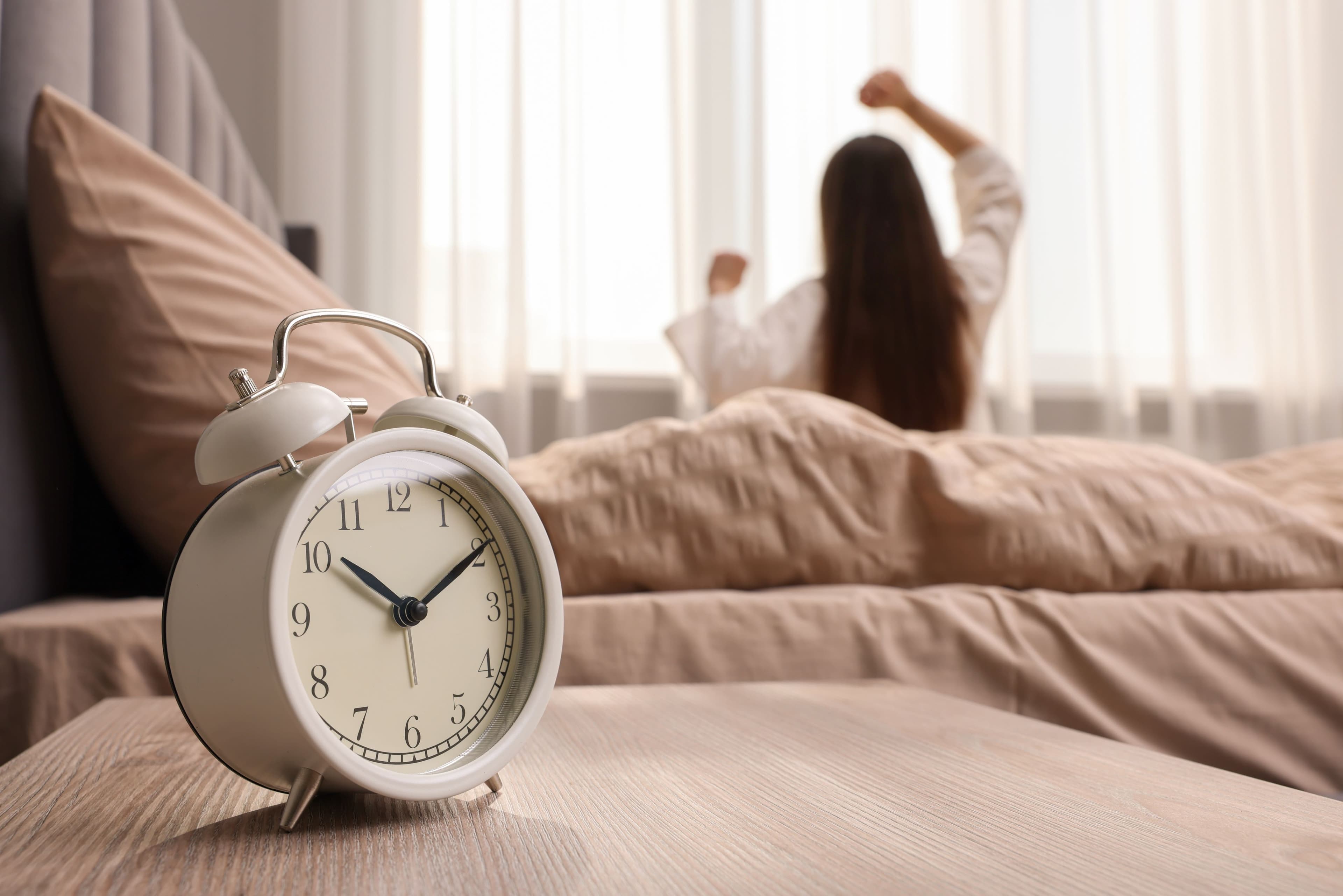 Alarm clock on a bedside table with a woman stretching on the bed behind