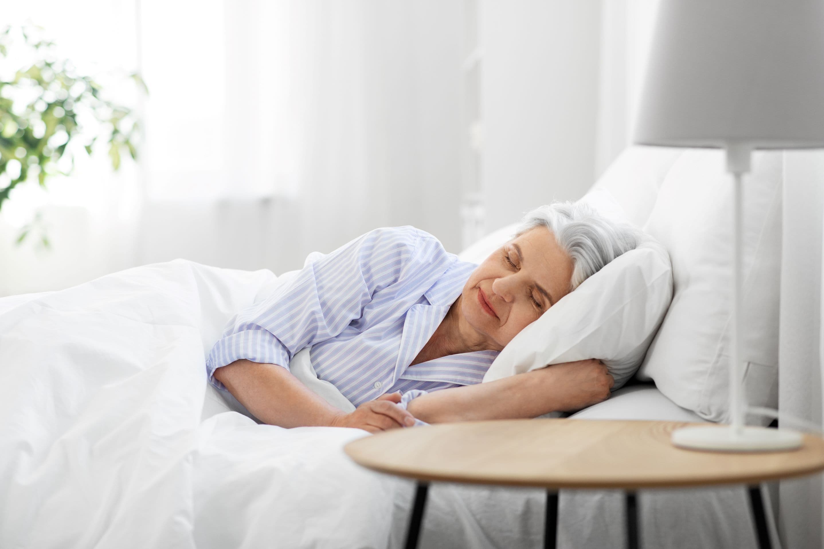Older woman with grey hair sleeping in a bright bedroom beside a lamp